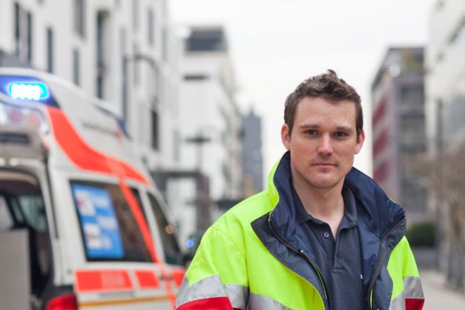 Foto: Ein junger DRK-Mitarbeiter steht mit einer Trage vor einem Einsatzfahrzeug mit eingeschaltetem Blaulicht.