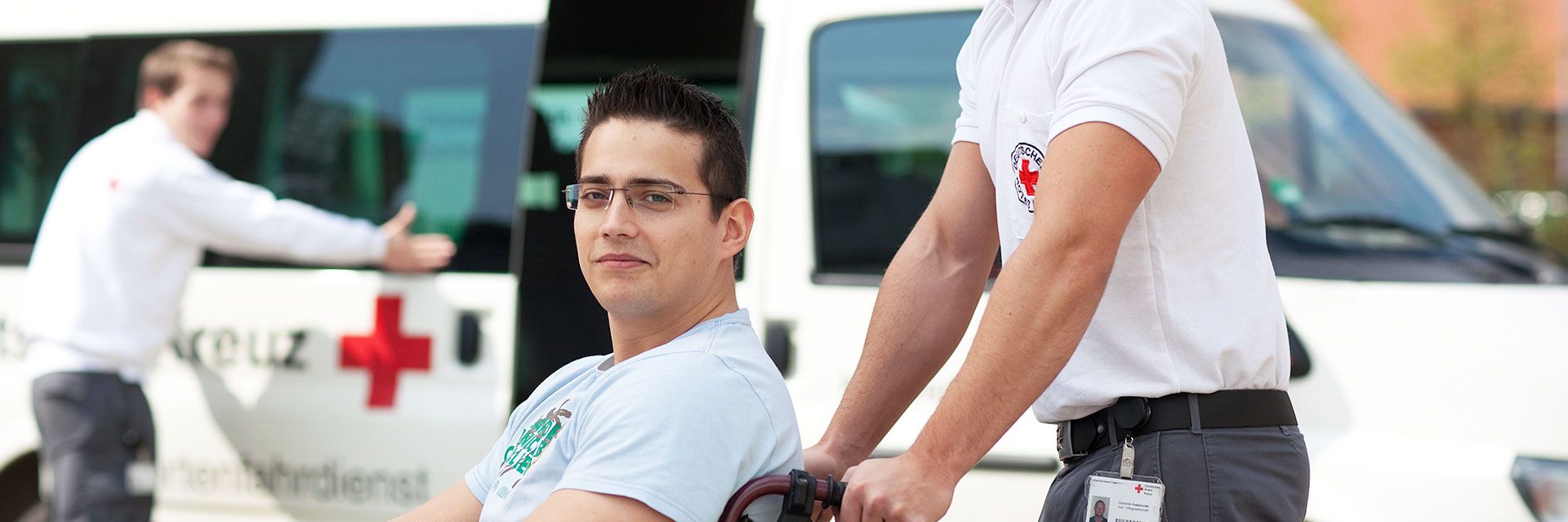 Foto: Ein junger Mann im Rollstuhl blickt zufrieden in die Kamera. Er wird von einem Mitarbeiter des DRK-Fahrdientes geschoben. Im Hintergrund öffnet ein weiterer Mitarbeiter die Schiebetür eines Transporters.